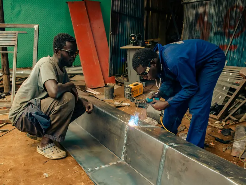 Two workers welding a long stainless steel channel in a fabrication yard